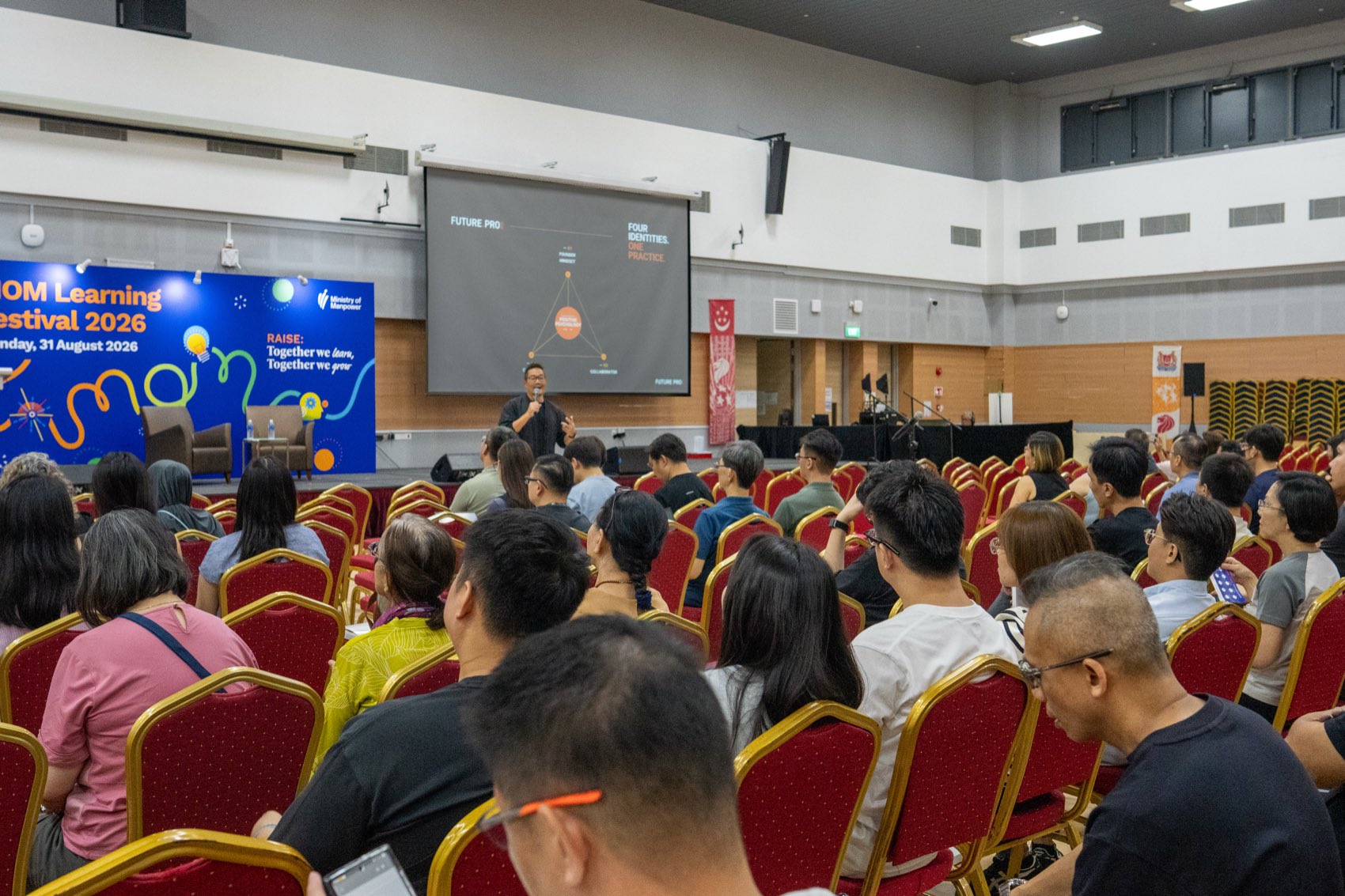 A full auditorium at a learning festival, with the four identities on screen behind the facilitator.